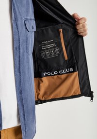 Hand som håller en öppen svart och brun Polo Club-jacka som visar innerfickan, loggan och etiketter för högkvalitativa ytterplagg.