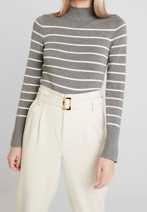Kvinde iført en grå og hvid stribet langærmet sweater og cremefarvede bukser med høj talje, bredt bælte og rektangulært spænde.
