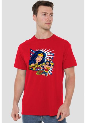 Mann trägt rotes Wonder Woman T-Shirt mit Comic-Stil-Grafik und Text, blickt nach rechts vor hellem einfarbigem Hintergrund.