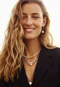 Lagdelte smykker med et design af sølvkæder, hvide perler og uregelmæssige sølvaccenter. Bår over en mørk blazer med dyb udskæring.