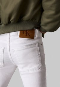 Witte denim jeans met een leren Calvin Klein Jeans-label op de achterkant van de taille. De outfit wordt aangevuld met een donkergroene bomberjack.