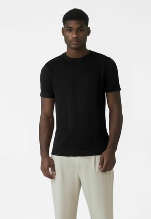 Sort striktrøje med korte ærmer, tekstureret mønster og rund halsudskæring, stylet med lyse bukser. Ingen synlige logoer eller grafik.