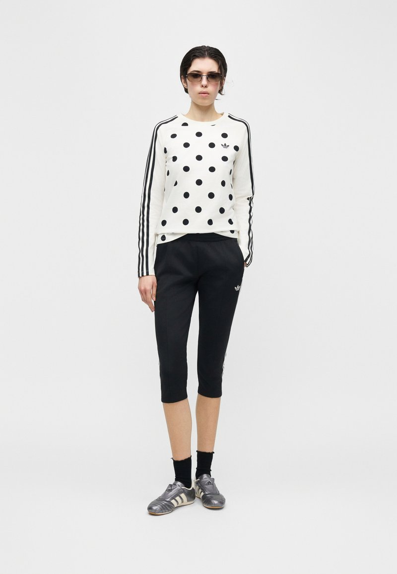 Kvinde iført hvid langærmet bluse med prikker, sorte korte bukser, sorte sokker, sølvfarvede sneakers og solbriller, stående mod en ensfarvet baggrund.