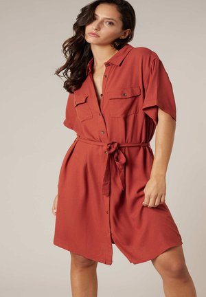 Femme portant une robe-chemise ceinturée de couleur rouille à manches courtes avec des poches poitrine sur un fond neutre.