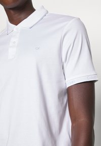 Vit pikétröja i bomull med en tre-knappars knappslå och en subtil CK-logotyp. Manschetterna har en ljusgrå Calvin Klein-detalj.