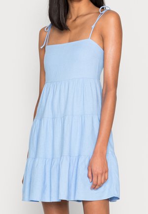 Femme portant une robe légère bleu clair à volants sans manches avec de fines bretelles nouées aux épaules, debout devant un fond neutre.