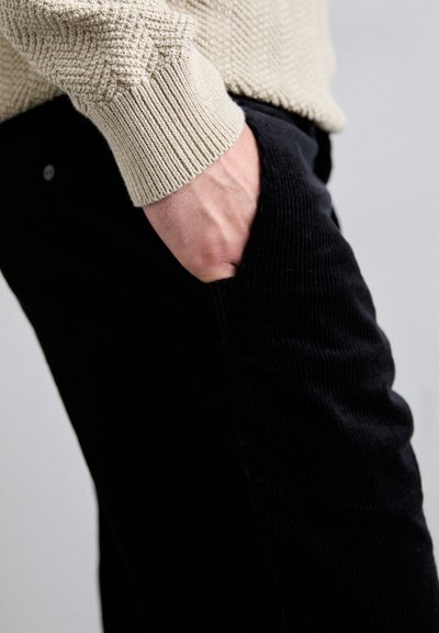 Pull en maille beige avec un motif en chevrons texturé, associé à un pantalon en velours côtelé noir. Main dans la poche, mettant en avant les détails du tissu.
