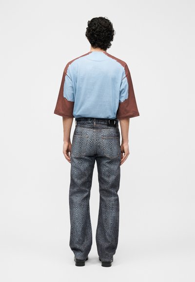 Άνδρας με σγουρά σκουρόχρωμα μαλλιά στέκεται με την πλάτη, φοράει άνετο μπλε και καφέ κοντομάνικο πουκάμισο και γκρι παντελόνι με τύπωμα λεοπάρδαλης, με μαύρα παπούτσια.