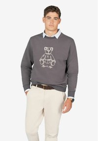 Grijze sweatshirt met een witte omlijnde teddybeertje ontwerp en tekst eronder. Gedragen over een lichtblauw overhemd met kraag, gecombineerd met crèmekleurige broek.