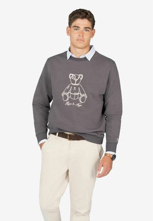 Sudadera gris con un diseño de oso de peluche contorneado en blanco y texto debajo. Usada sobre una camisa de cuello azul claro, combinada con pantalones color crema.