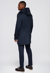 Abrigo aislante azul marino con capucha, que presenta un corte ajustado, bolsillos laterales y puños abotonados. Llevado sobre pantalones y botas a juego.