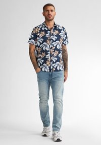 Camicia a maniche corte navy con motivi floreali bianchi e beige tropicali, abbinata a jeans azzurri e sneakers bianche.