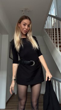 Zwarte jurk met lange mouwen en een aansluitende silhouet, voorzien van een brede riem. Doorzichtige zwarte panty's en een zwarte jas in één hand gehouden. Licht houten vloer.