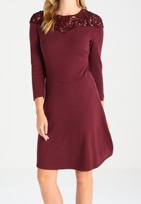 Mujer vistiendo un vestido burdeos de manga larga y a la altura de la rodilla, con detalles de encaje en el pecho y los hombros.