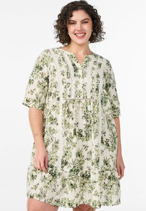 Uśmiechnięta kobieta o kręconych włosach ubrana w kremową sukienkę do kolan z zielonym kwiatowym wzorem i koronkowymi detalami, stojąca na białym tle.