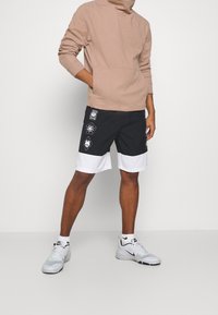 Pantalones cortos negros con dobladillo blanco, con estampados gráficos en blanco. Combinados con una sudadera color marrón claro con un bolsillo frontal, usados con zapatillas blancas.