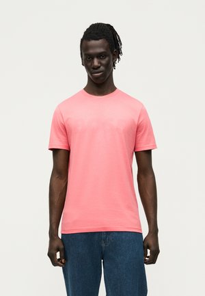 Jeune homme à la peau foncée avec des dreadlocks, portant un t-shirt rose uni et un jean bleu, debout devant un fond clair.