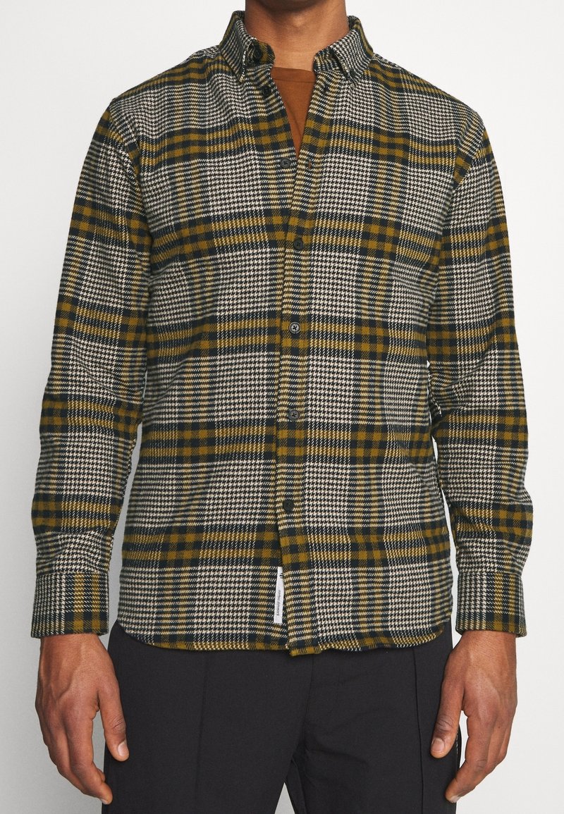 Homme portant une chemise à manches longues à carreaux noirs, blancs et vert olive par-dessus un sous-vêtement marron, pantalon noir.