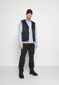 Chaleco acolchado negro sobre sudadera azul clara con logo, pantalones cargo negros y zapatillas negras. Se aprecian líneas limpias y bolsillos utilitarios.