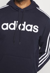 Sweat à capuche bleu marine avec le logo "adidas" blanc, doté d'une poche kangourou, d'un cordon de serrage réglable et de rayures blanches sur les manches.