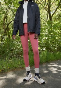 Veste imperméable noire par-dessus un haut zippé blanc, associée à des leggings roses et des chaussettes blanches. Les chaussures présentent un design noir et crème, avec des accents orange.
