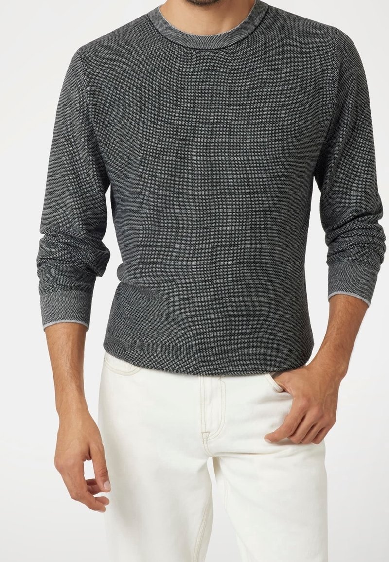 Homme portant un pull à manches longues texturé gris foncé et un pantalon blanc, une main dans la poche et l'autre détendue le long du corps.