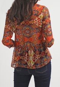 Blusa colorata e decorata in tessuto trasparente con vita a peplum, caratterizzata da vivaci tonalità di rosso, arancione e blu. Maniche lunghe con polsini bottonati.