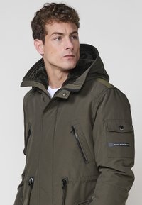 Tumši zaļš parka jakas modelis ar noņemamu kapuci, mākslīgo kažokādas oderi, ar rāvējslēdzēju kabatām un siksnu akcentiem uz piedurknēm, nodrošinot funkcionālu dizainu.