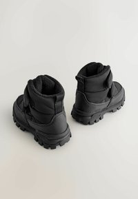 Sorte isolerede støvler med polstret design, velcrolukning og teksturerede gummisåler for godt greb. Har en blød foring og solid konstruktion.