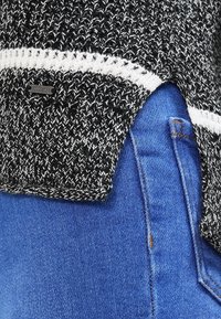 Neule mustavalkoinen villapaita, jossa on tekstuurinen kangas ja sivuaukko, yhdistettynä sinisiin farkkuihin, jotka esittelevät klassista istuvuutta ja ompelua.