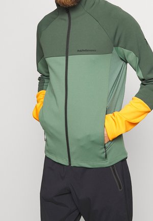 Veste zippée verte et orange avec un avant texturé, des manches lisses, des poches latérales et des panneaux contrastants. Muni du logo 'Peak Performance'.