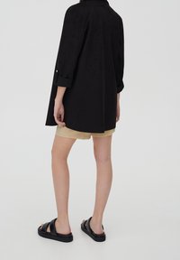 Camisa de ante negro con cuello y mangas enrolladas, combinada con pantalones cortos beige. Sandalias negras con tiras anchas complementan el conjunto.