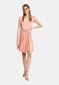 Abito corto rosa con scollatura quadrata, maniche a cappuccio e gonna plissettata. Gli accessori in oro in vita aggiungono dettagli. Tessuto dalla texture liscia.