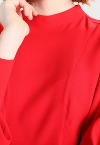 Rød bluse med høj hals lavet af glat stof. Har lange ærmer og diskrete syninger. Close-up af halsudskæringen og skulderområdet.
