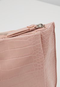 Gros plan d’un sac en cuir rose à motif crocodile avec une fermeture éclair argentée partiellement ouverte sur un fond uni.