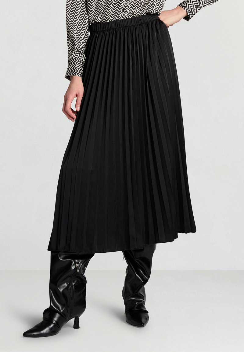 Donna che indossa una gonna midi plissettata nera, stivali neri alti fino al ginocchio con tacco e una maglietta a maniche lunghe con motivo bianco e nero.