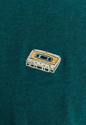 Cinta de casete bordada sobre un fondo de tela verde azulado, con detalles en beige y negro, forma rectangular y punto texturizado.