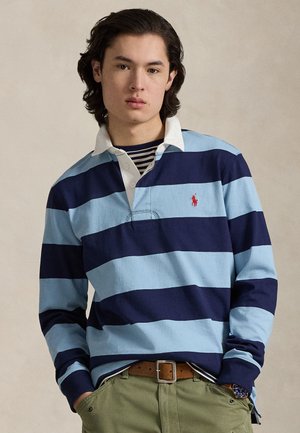 Długi rękaw w jasnoniebieskie i granatowe paski polo z białym kołnierzykiem i haftowanym logo. Noszona na granatowo-białej koszulce w paski.