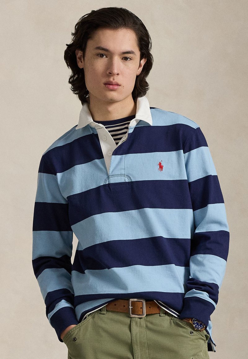 Polo a maniche lunghe a righe azzurro chiaro e blu navy con colletto bianco e logo ricamato. Indossata sopra una maglietta a righe blu navy e bianche.