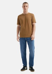 Brązowa bawełniana koszulka, krótki rękaw, okrągły dekolt, w połączeniu z niebieskimi dżinsami. Czarne sneakersy z brązowymi podeszwami dopełniają strój.