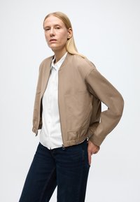 Vajszínű bomber dzseki cipzáras záródással, rugalmas mandzsettákkal és elülső zsebbel, fehér gombos ing és sötét farmer fölött viselve. Sima textúra.