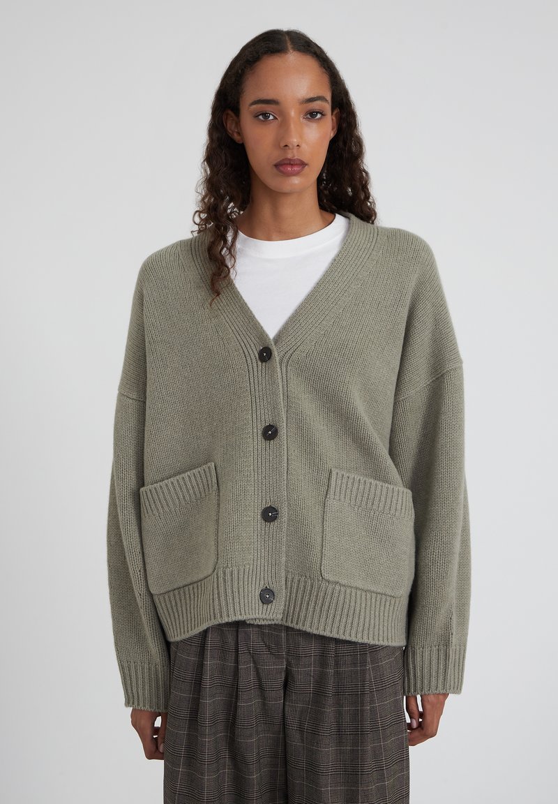 Femme portant un cardigan gris clair surdimensionné avec des boutons noirs, par-dessus une chemise blanche et un pantalon à carreaux, debout devant un fond uni.