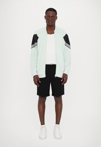 Casaco atlético verde-menta com detalhes e riscas pretas, usado sobre uma camisa branca e combinado com calções pretos e ténis brancos.