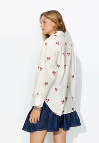 Chemise blanche à manches longues avec broderie florale rouge, coupe décontractée, col boutonné. Associée à une jupe évasée en denim foncé.
