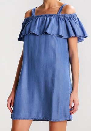 Vrouw draagt een mouwloze blauwe off-shoulder jurk met ruches over de borst en dunne schouderbandjes.