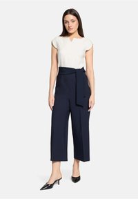 Blouse blanche à manches courtes avec un col en V et culottes taille haute bleu marine, ornées d'une ceinture nouée et de jambes larges. Escarpins noirs à bout pointu.