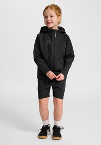 Svart huva jacka med dragkedja, i kombination med svarta shorts. Båda har en slät textur. Innehåller logotyp på bröstet och atletiska skor.