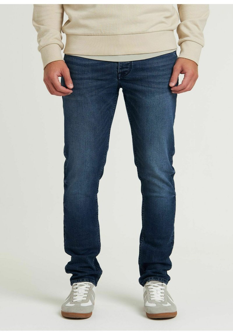 Jeans blu scuro con vestibilità slim, caratterizzati da un classico design a cinque tasche e leggere scoloriture. Indossati con sneakers grigie e bianche.