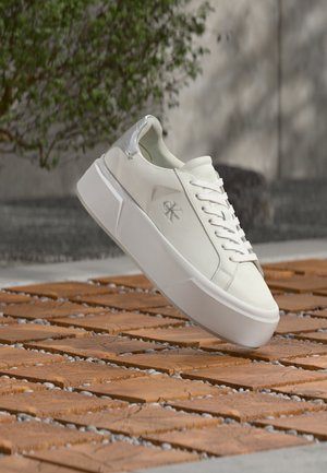 Λευκό αθλητικό παπούτσι Calvin Klein με χοντρή σόλα που αιωρείται πάνω από σχέδιο εξωτερικού χώρου με πράσινη βλάστηση στο φόντο.