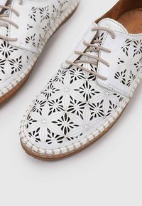 Zapatos de cuero blanco con un diseño de corte floral, punta redonda, cordones beige y una costura intrincada a lo largo de la suela.